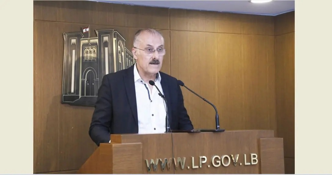 لقطة متوسطة للنائب بلال عبد الله مرتدياً بدلة داكنة وهو يلقي كلمة خلف منصة خشبية تحمل شعار ورابط موقع البرلمان اللبناني.