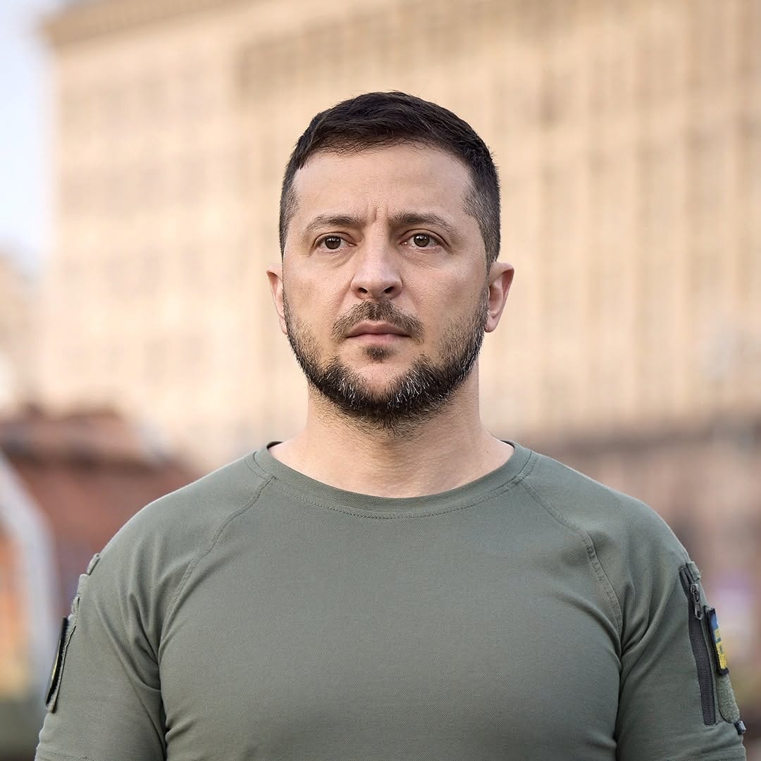Close-up portrait of Ukrainian President Volodymyr Zelenskyy wearing an olive green t-shirt, standing outdoors with a serious expression.